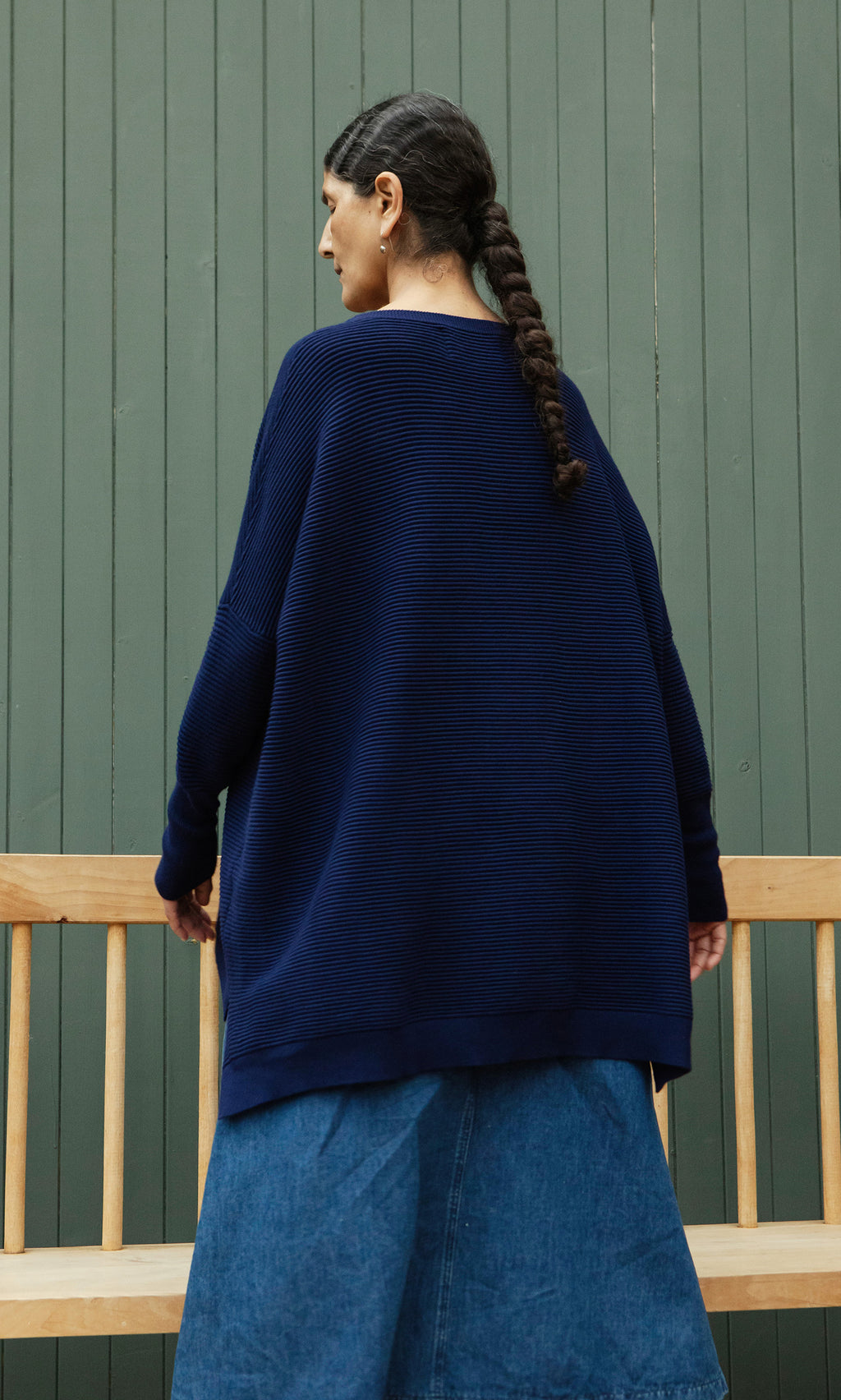Plümo  Woman with a long braid wears the Jack Sweater - Navy, oversized with dropped shoulders, and a denim skirt by a bench.