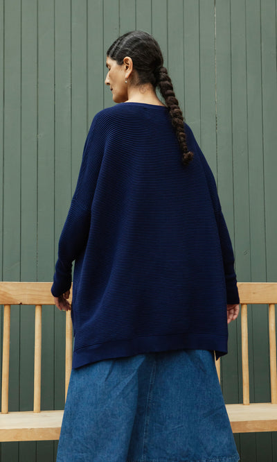 Plümo  Woman with a long braid wears the Jack Sweater - Navy, oversized with dropped shoulders, and a denim skirt by a bench.
