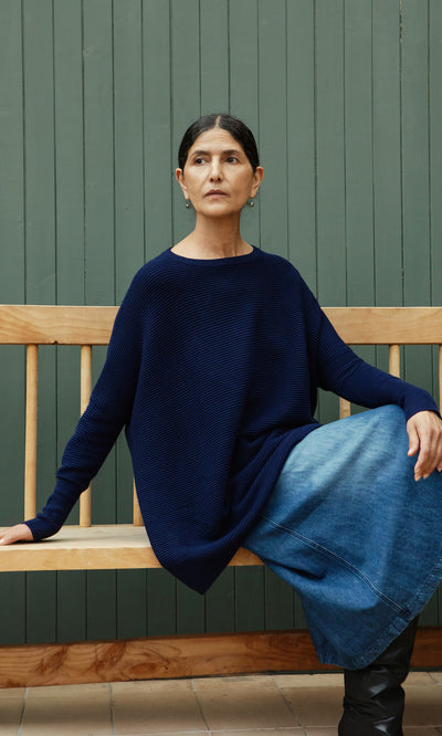 Plümo  Woman in the Jack Sweater - Navy and a denim skirt sits on a bench by a green paneled wall.
