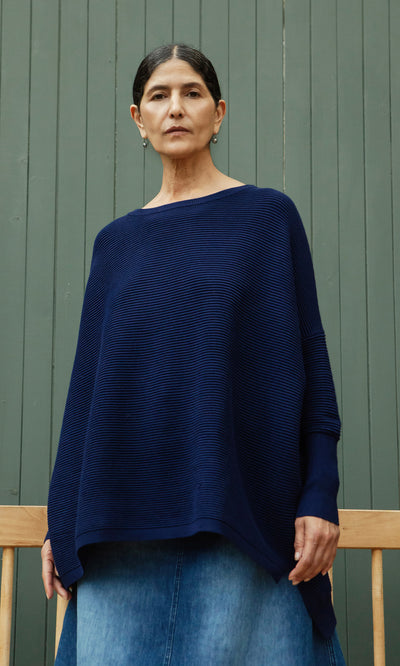 Plümo  Woman in the Jack Sweater - Navy with dropped shoulders and a denim skirt stands before a green wooden background.
