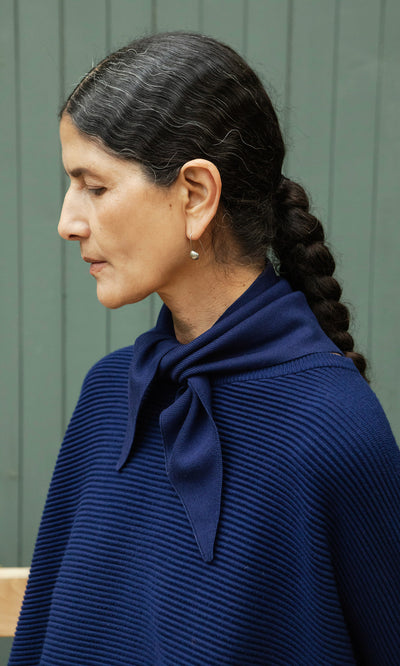 Plümo  Woman with dark braids in a navy Jona Necktie, standing in profile against a green wall.
