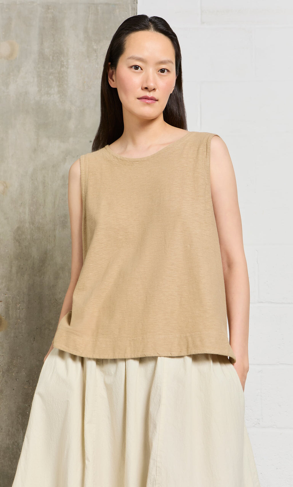 Plümo- Woman wearing the Jersey Top, a loose beige tank, against a neutral textured background. Sustainably produced.