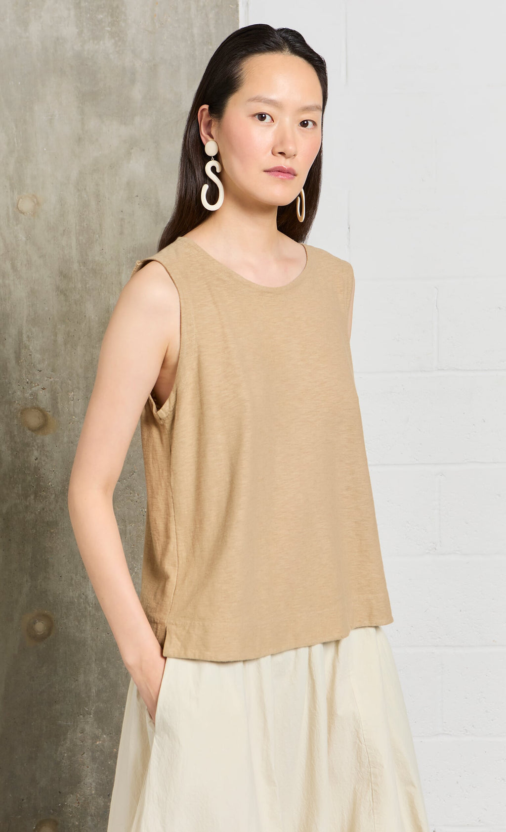 Plümo- Woman wearing Horn Clip-On Earrings, a sleeveless beige top, and cream skirt, standing by a textured wall.