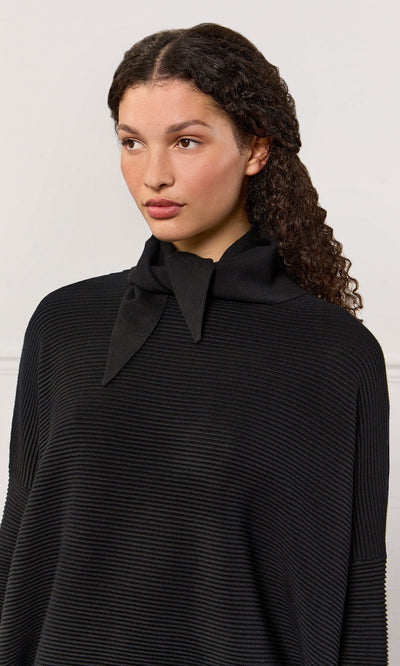 Plümo- A woman with curly hair wears a black Jona Necktie top with a folded collar, looking slightly to the side.