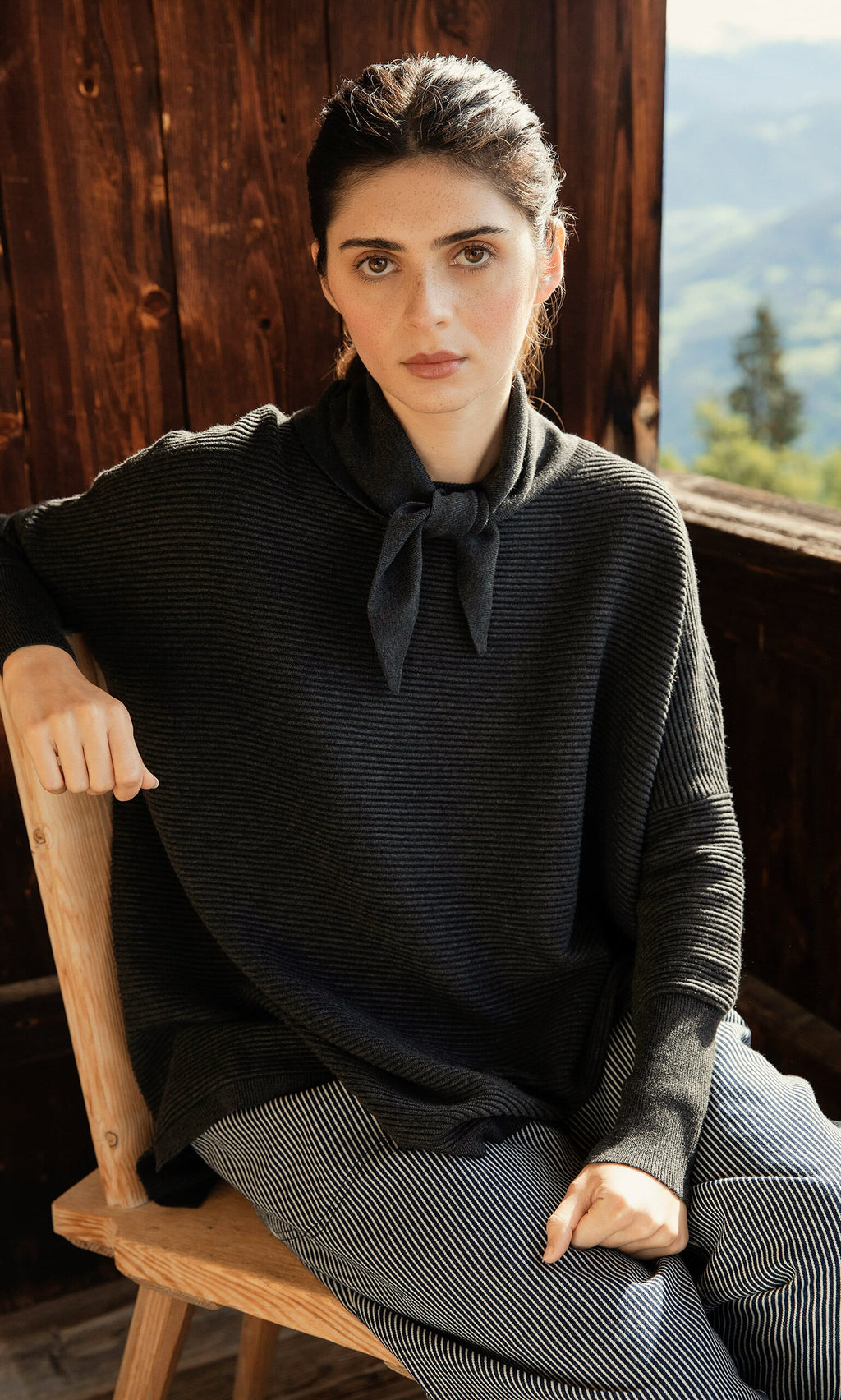 Plümo- Woman with dark hair wears the Jona Necktie in Graphite, sitting on a wooden chair outdoors.