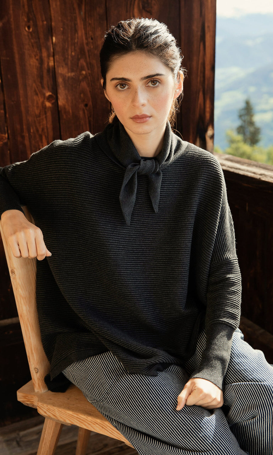 Plümo- Woman with dark hair wears the Jona Necktie in Graphite, sitting on a wooden chair outdoors.