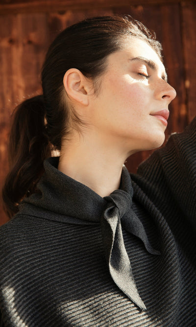 Plümo- Woman with dark hair in a ponytail, eyes closed in sunlight, wears the Jona Necktie - Graphite top.
