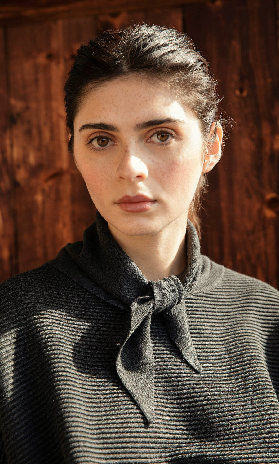 Plümo- Woman with dark hair wears the Jona Necktie – Graphite top, standing before a wooden background.