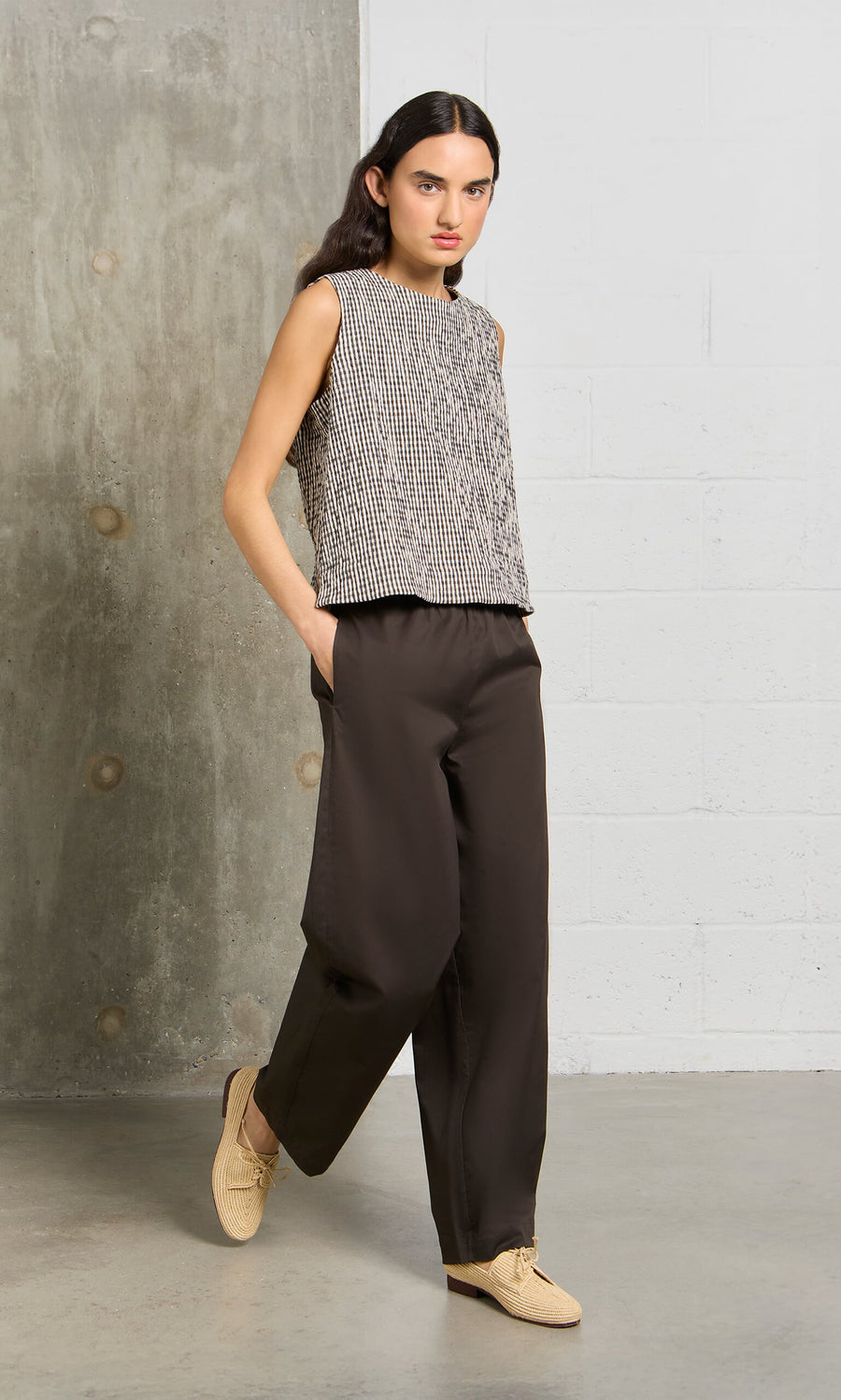 Plümo- Woman in the Linda Top - Gingham, dark wide-leg pants, and beige shoes against a concrete and white wall.