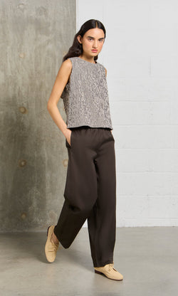 Plümo- Woman in a sleeveless patterned top, Kathleen Pants in organic cotton, and beige shoes stands by a concrete wall.