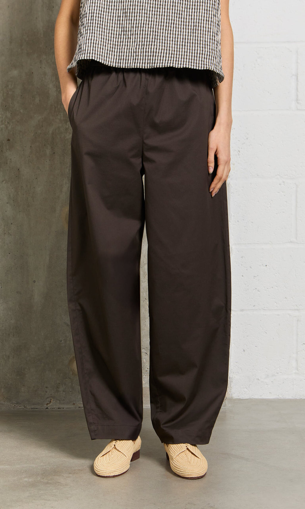 Plümo- Person wears Kathleen Pants, beige shoes, and a checkered top while standing by a concrete and white wall.