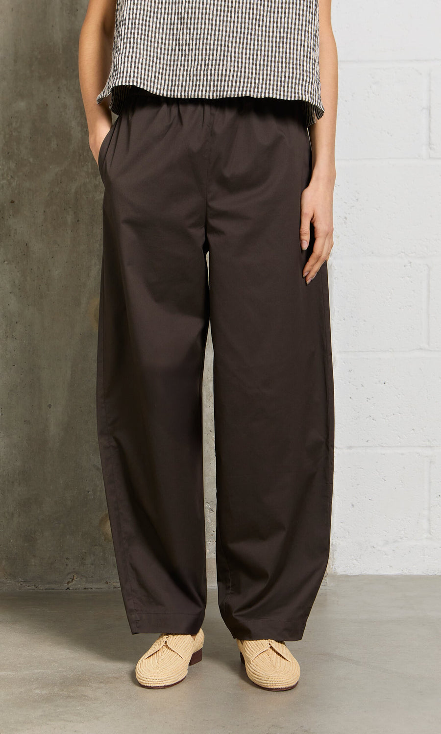 Plümo- Person wears Kathleen Pants, beige shoes, and a checkered top while standing by a concrete and white wall.
