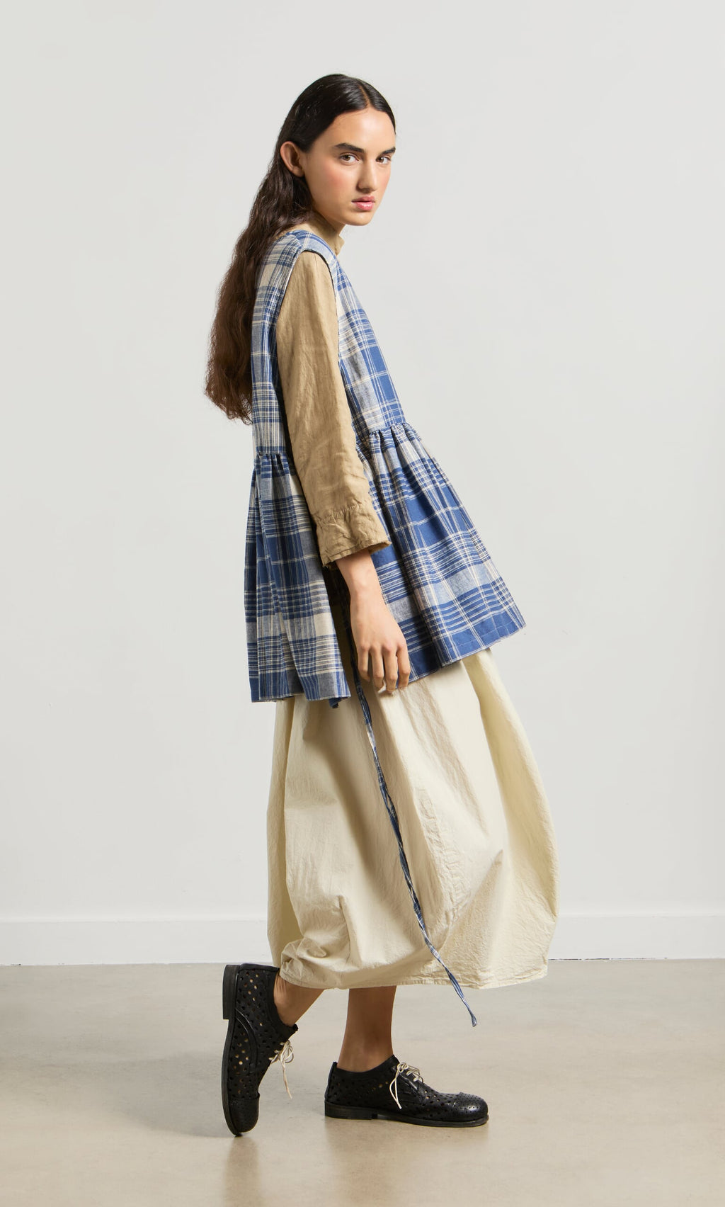 Plümo- Person in the Keefe Tabbard—a blue plaid top—over a tan dress and black shoes stands sideways against a plain background.