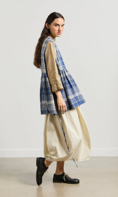 Plümo- Person in the Keefe Tabbard—a blue plaid top—over a tan dress and black shoes stands sideways against a plain background.