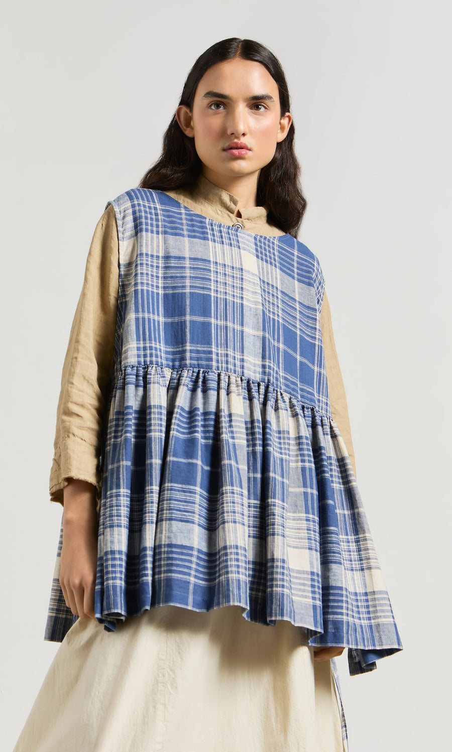 Plümo- Person wearing the Keefe Tabbard in blue-and-white plaid over a beige shirt and cream skirt, against a plain background.