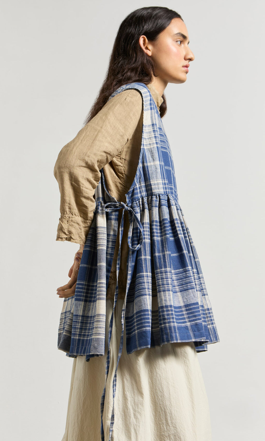 Plümo- Woman in profile wearing the Keefe Tabbard over a cream skirt, styled with a blue plaid sleeveless dress tied at the side.