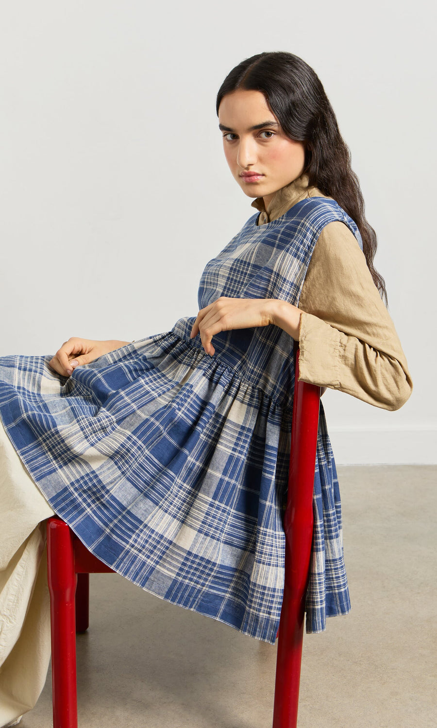 Plümo- A woman in the Keefe Tabbard sits sideways on a red chair, looking at the camera with a neutral expression.