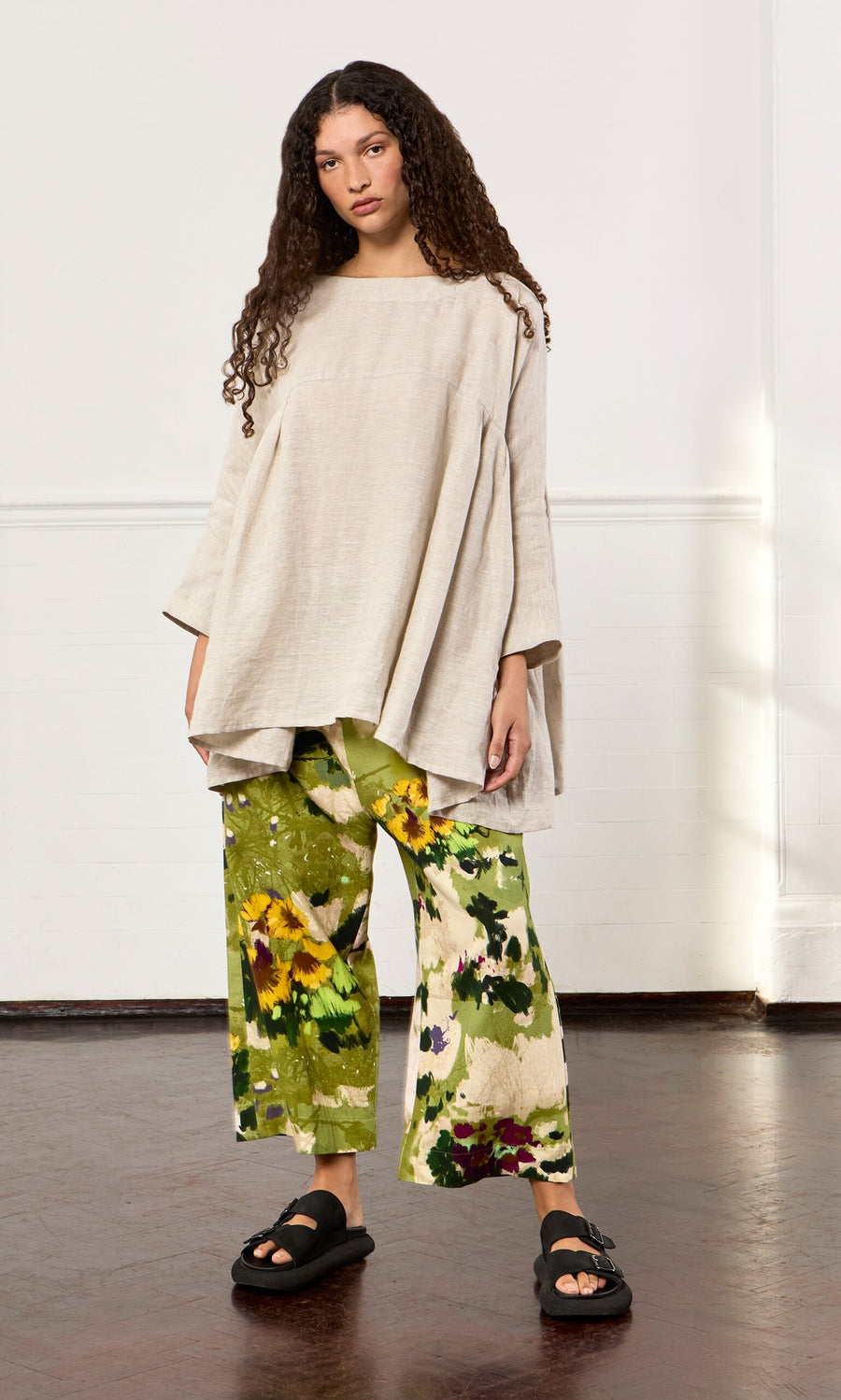 Plümo- A woman in the Kelli Blouse, floral green pants, and black sandals stands against a white wall.