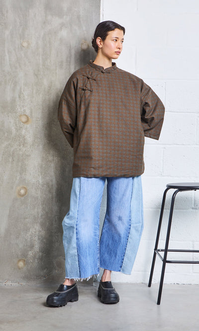 Plümo- Person in a Kenza Padded Top and wide, two-tone jeans stands by a concrete wall next to a black stool.