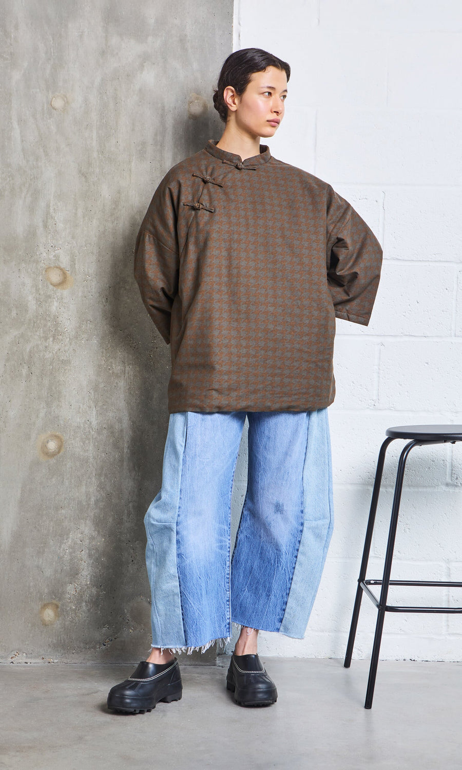 Plümo- Person in a Kenza Padded Top and wide, two-tone jeans stands by a concrete wall next to a black stool.