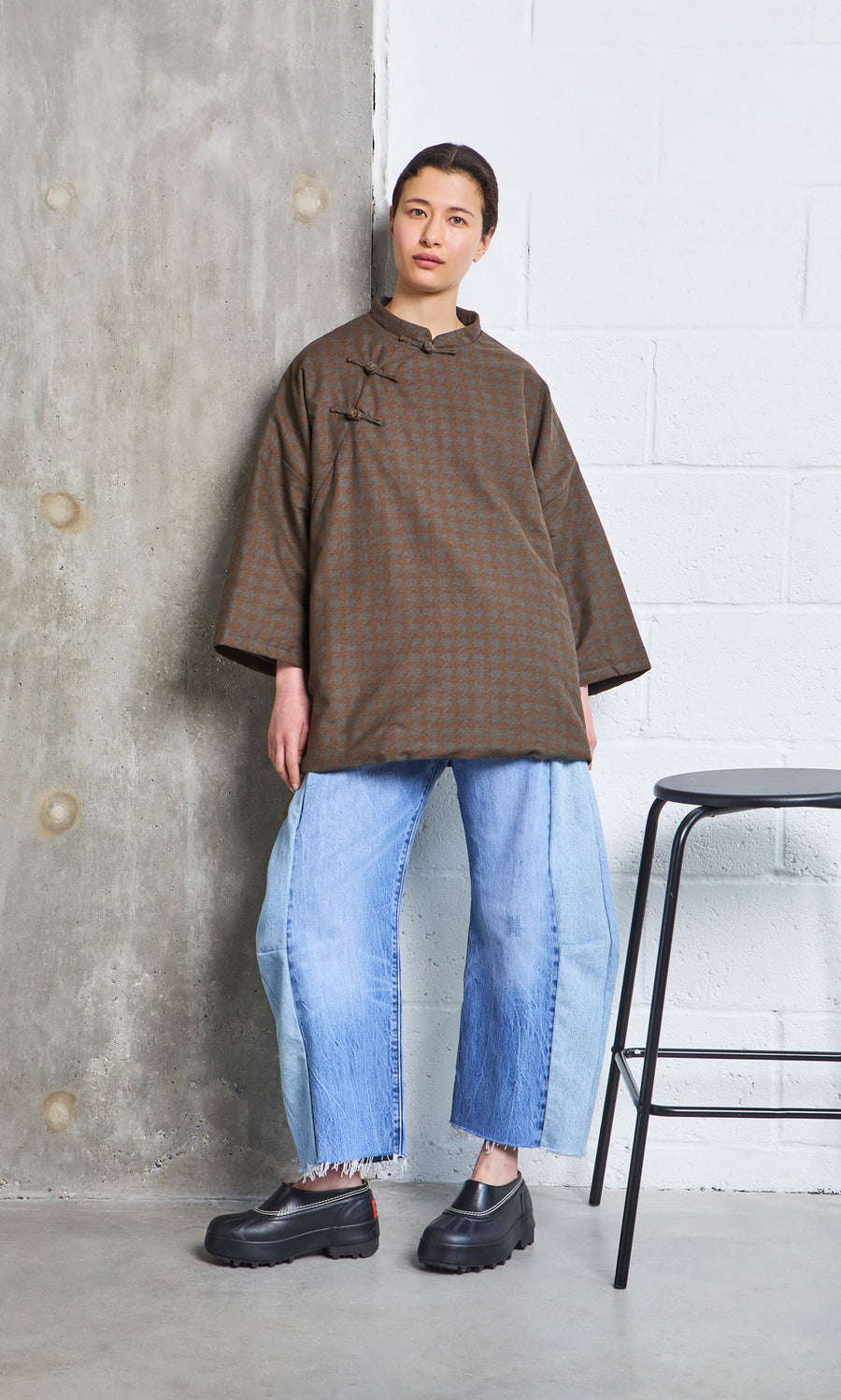 Plümo- A person in a Kenza Padded Top and wide, light blue jeans stands by a wall next to a black stool.