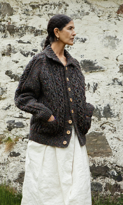 Plümo  Someone in a Kilcarra Tweed Cardigan and white skirt stands outside beside a textured stone wall.