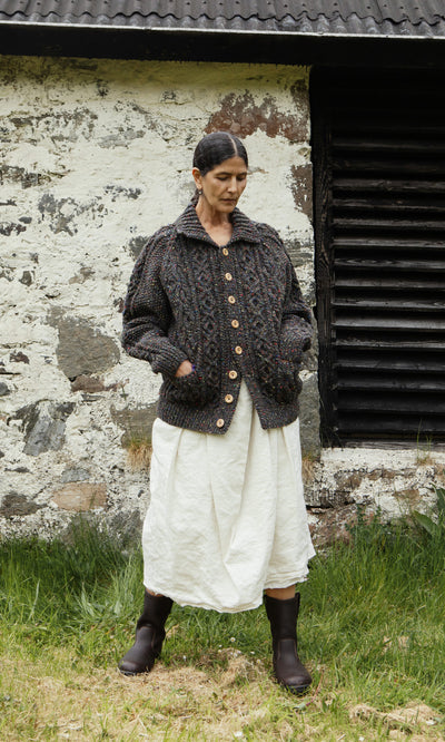 Plümo  A woman in a Kilcarra Tweed Cardigan, white skirt, and boots stands by a weathered stone wall and black window.