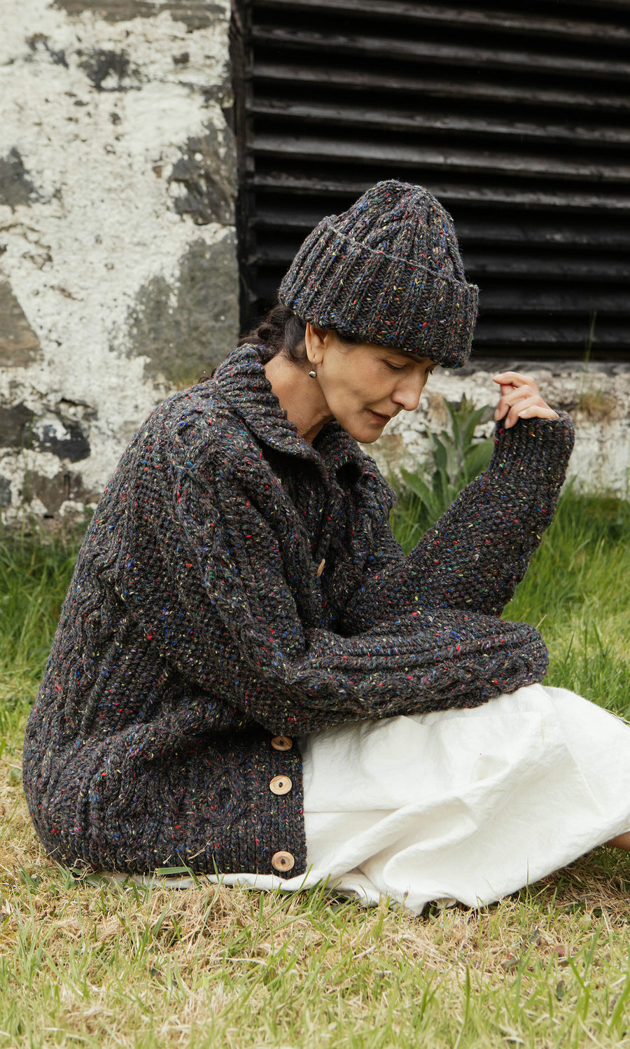 Plümo  Wearing a Kilcarra Tweed Beanie, someone sits on grass by a weathered wall, gazing downward softly.