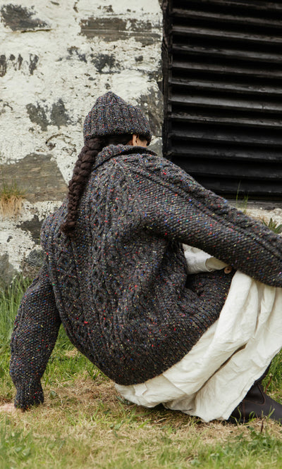 Plümo  Person in a millstone grey Kilcarra Tweed Cardigan and hat sits on grass, facing away near a weathered wall.