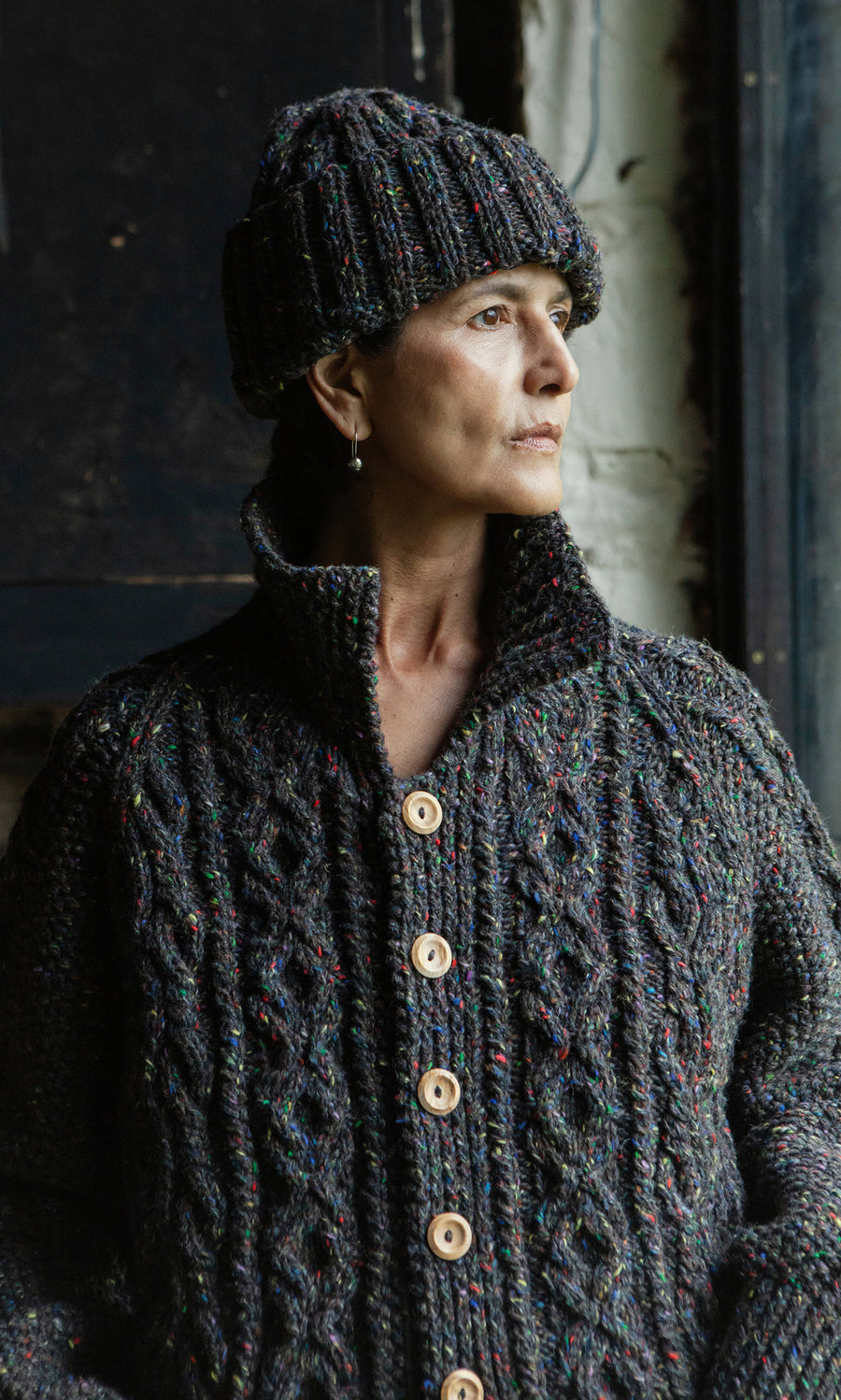 Plümo  Woman wearing a tweedy wool knit hat with a roll-up cuff and sweater, looking out a window thoughtfully.
