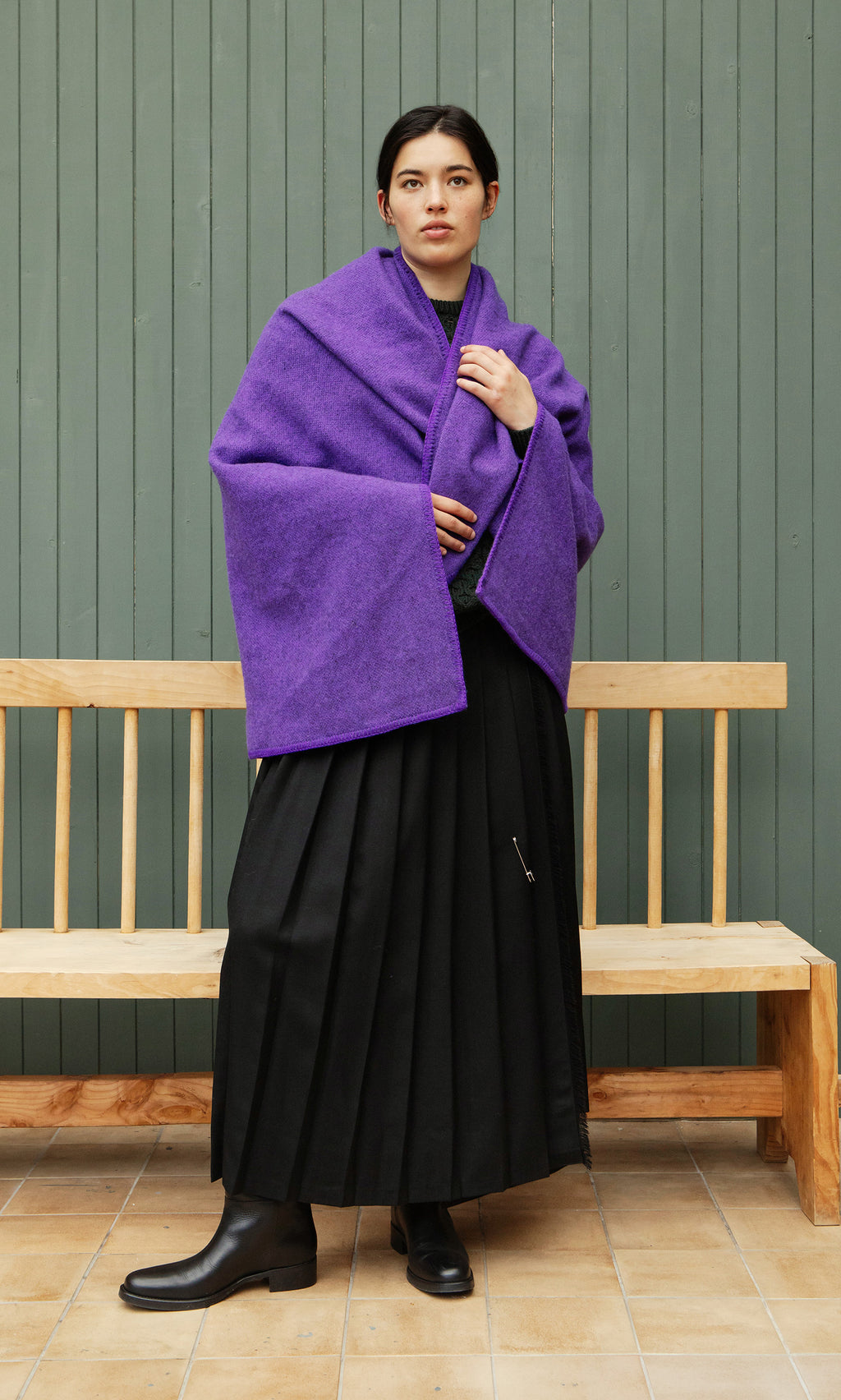 Plümo  A person in a Kilkenny Blanket - Violet and black skirt stands before a wooden bench and green paneled wall.