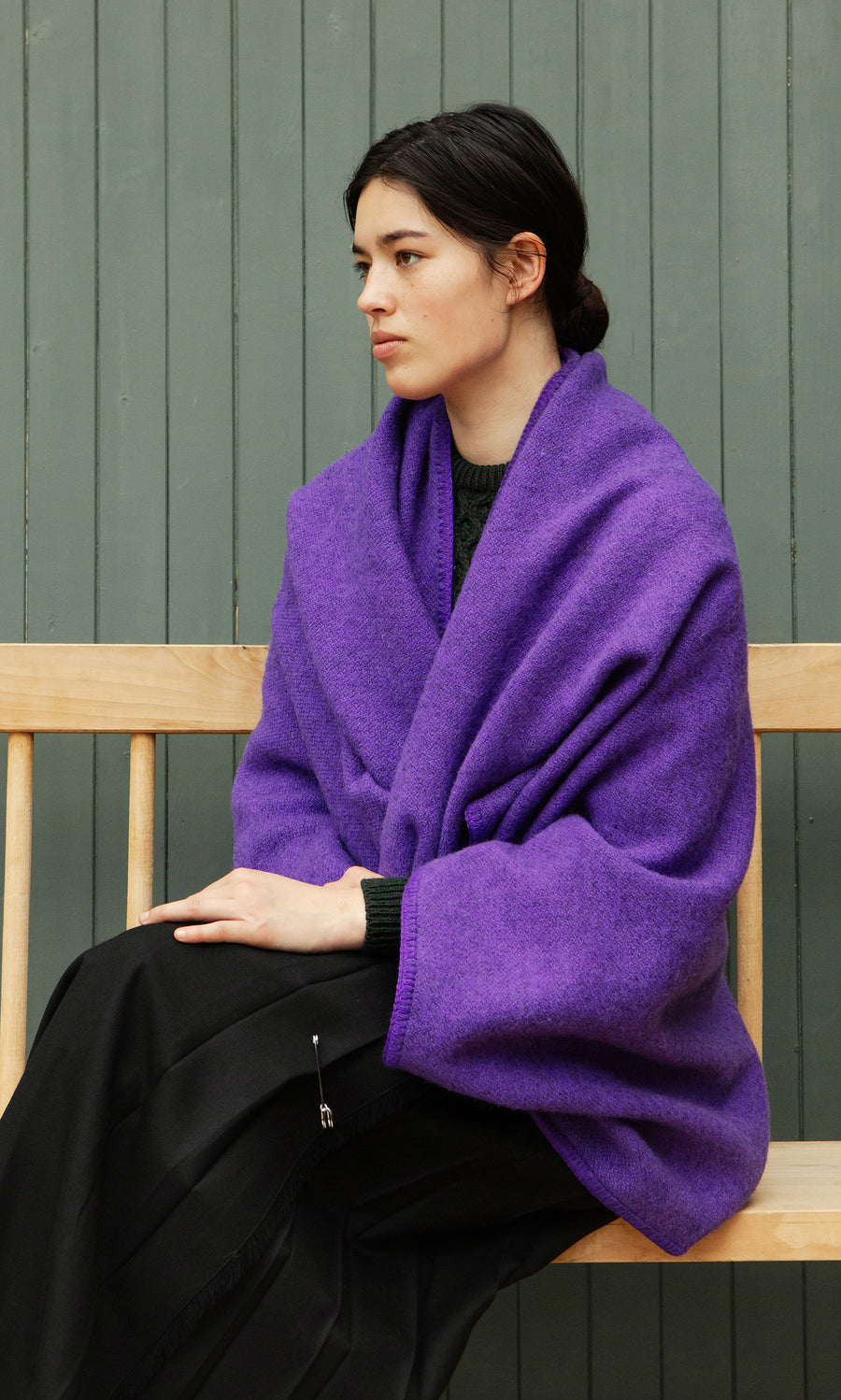 Plümo  Someone wrapped in a Kilkenny Blanket - Violet sits on a wooden bench by a green paneled wall, gazing sideways.