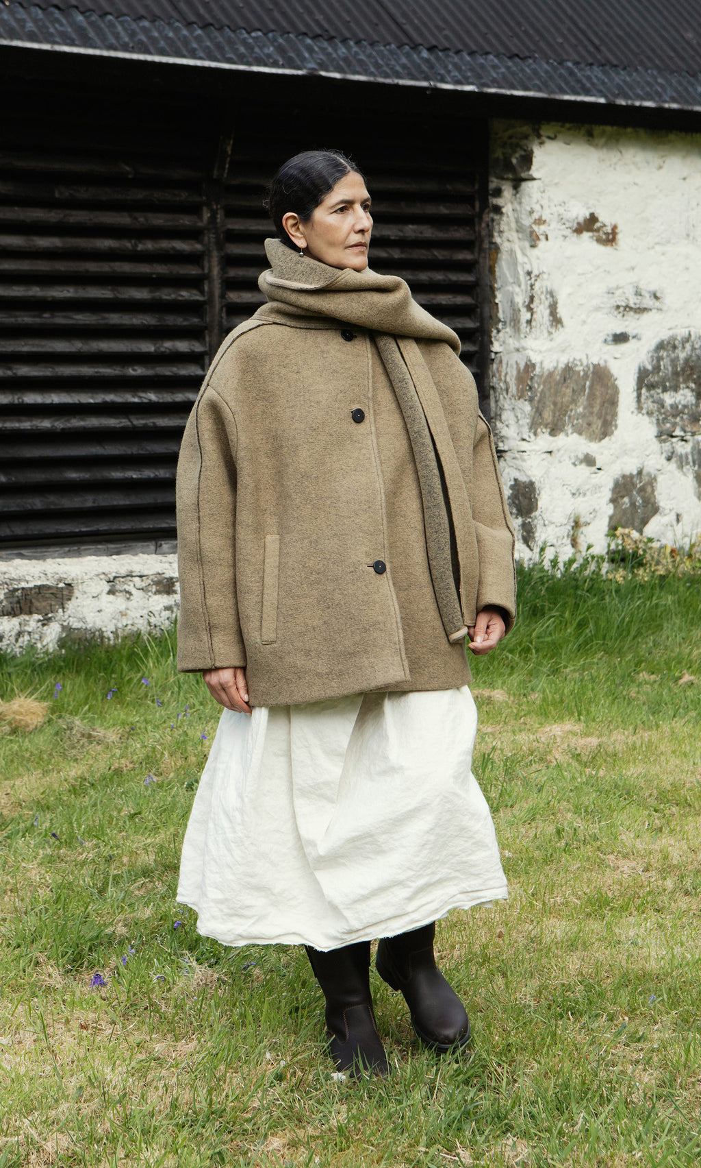 Plümo  Woman in a Kilmore Coat in acorn brown, white skirt, and boots stands on grass by a rustic stone building.
