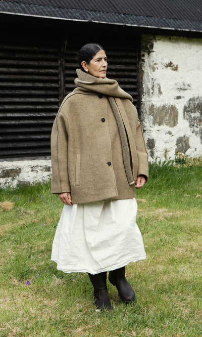Plümo  Woman in a Kilmore Coat in acorn brown, white skirt, and boots stands on grass by a rustic stone building.
