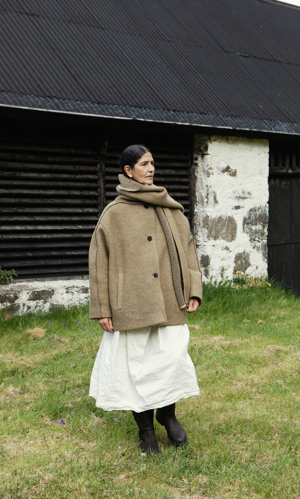 Plümo  Person in a Kilmore Coat with detachable scarf, white skirt, and boots stands on grass before a rustic building.