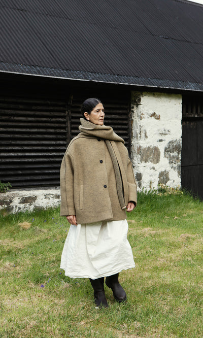 Plümo  Person in a Kilmore Coat with detachable scarf, white skirt, and boots stands on grass before a rustic building.