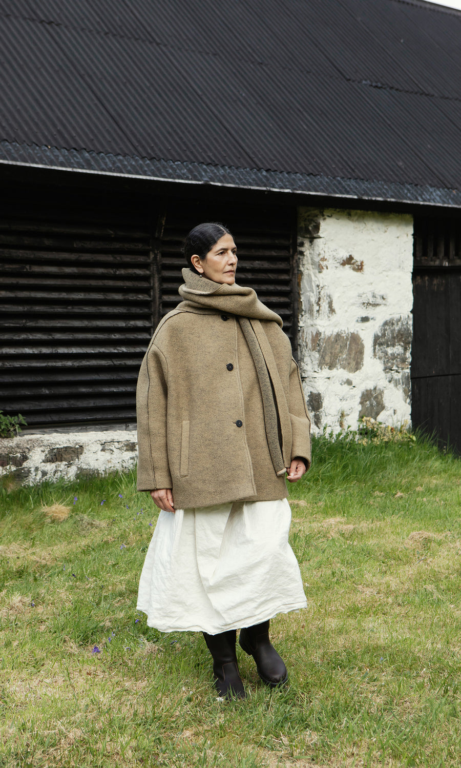 Plümo  Person in a Kilmore Coat with detachable scarf, white skirt, and boots stands on grass before a rustic building.
