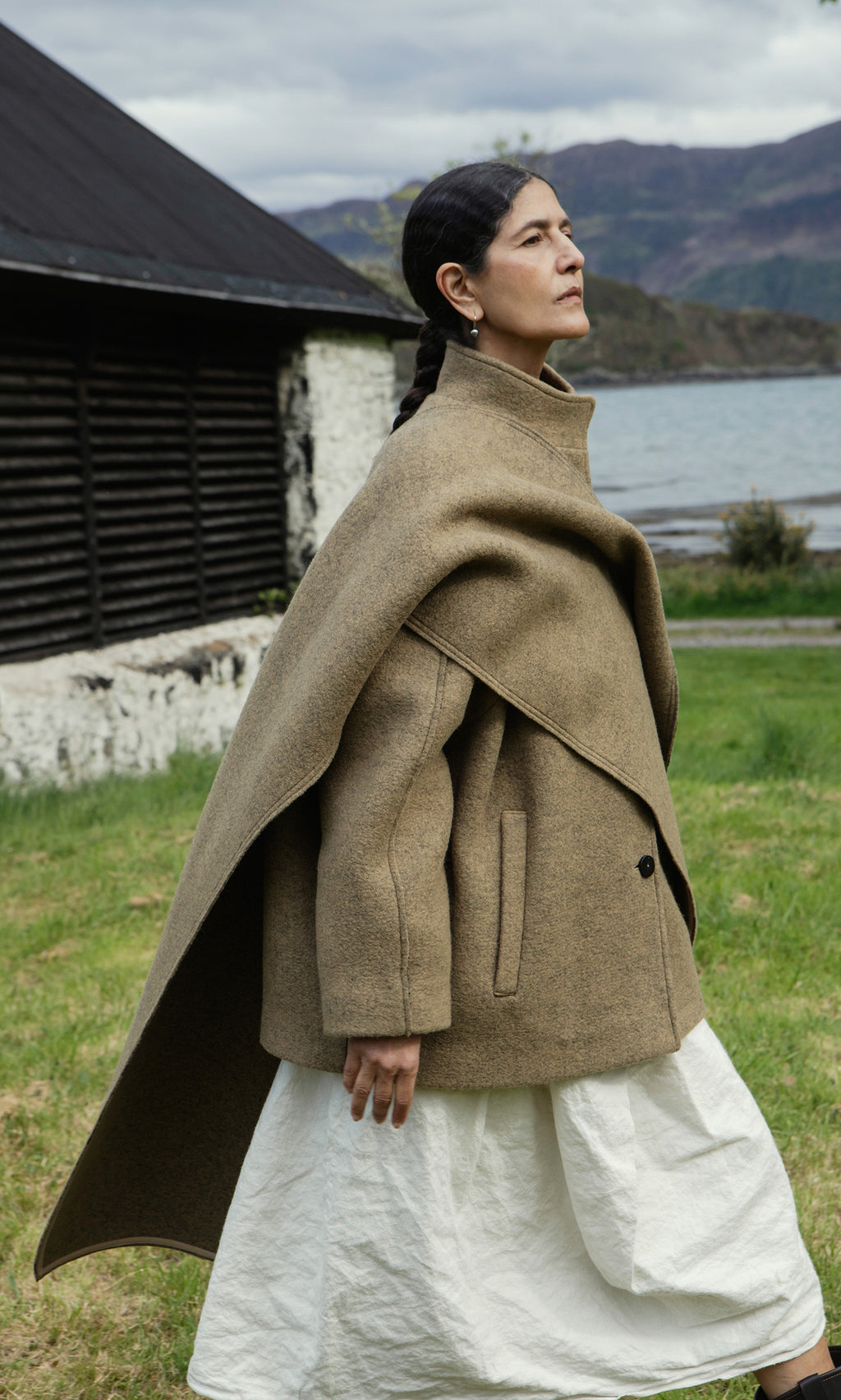 Plümo  Woman in a Kilmore Coat and white skirt walks by a rustic building and lake with mountains in the background.