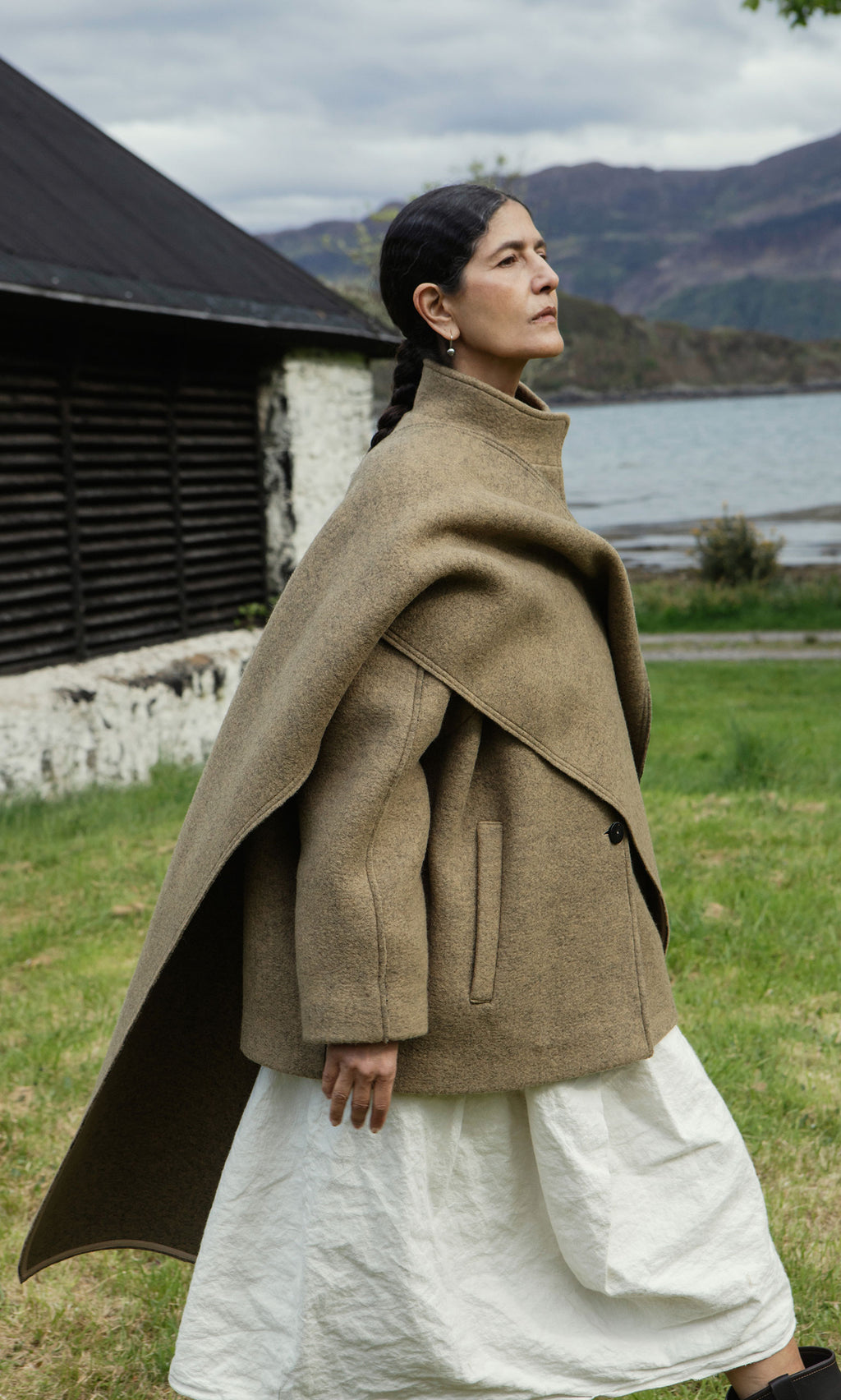 Plümo  A woman in a Kilmore Coat and white dress walks by a rustic lakeside building, mountains in the background.