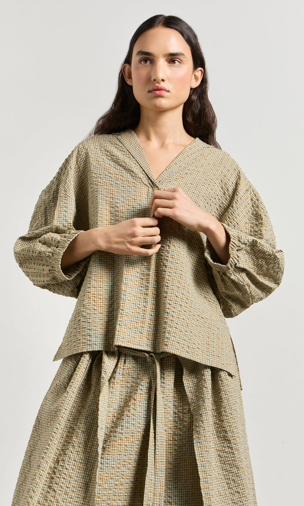 Plümo- Woman wearing the Fern Blouse, a boxy beige checkered cotton seersucker shirt made in London, against a light background.
