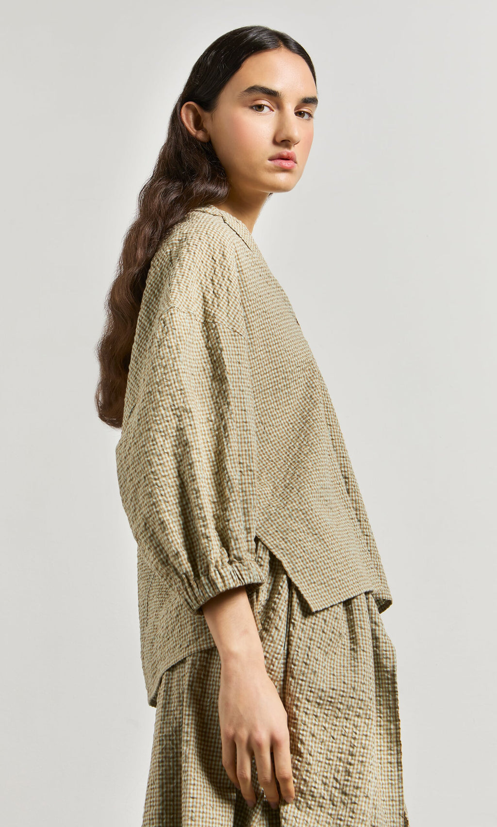Plümo- Person with long brown hair wears a made in London beige checked Fern Blouse, standing sideways against a light background.