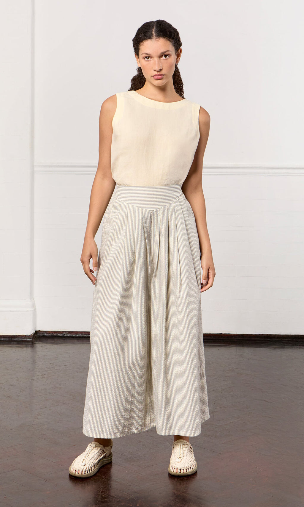 Plümo- A woman stands indoors in a sleeveless beige top, Laurel Pants with ticking stripes, and platform shoes.