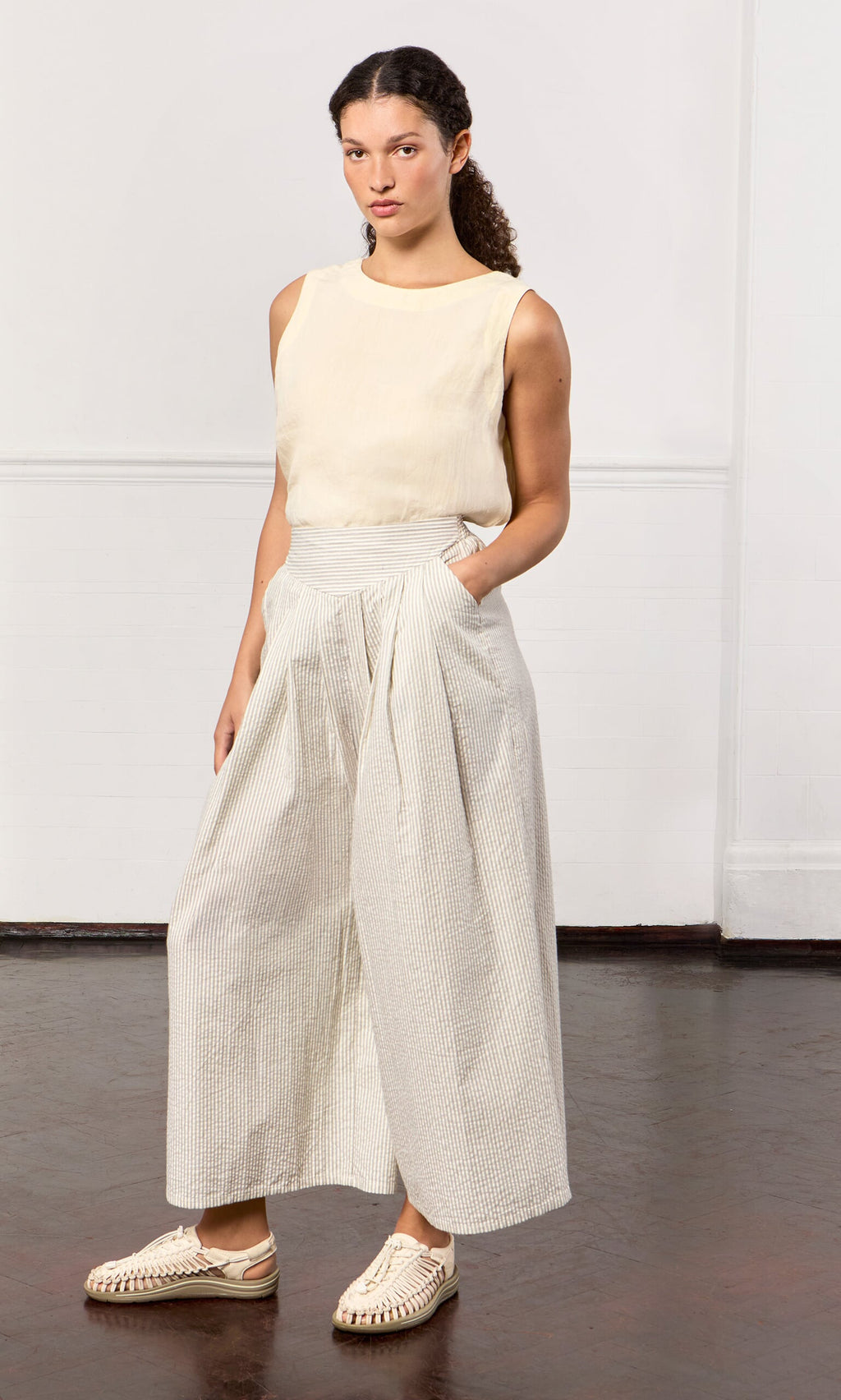 Plümo- Woman in Laurel Pants—ticking stripe cotton palazzo style with elastic waistband—paired with sleeveless beige top.
