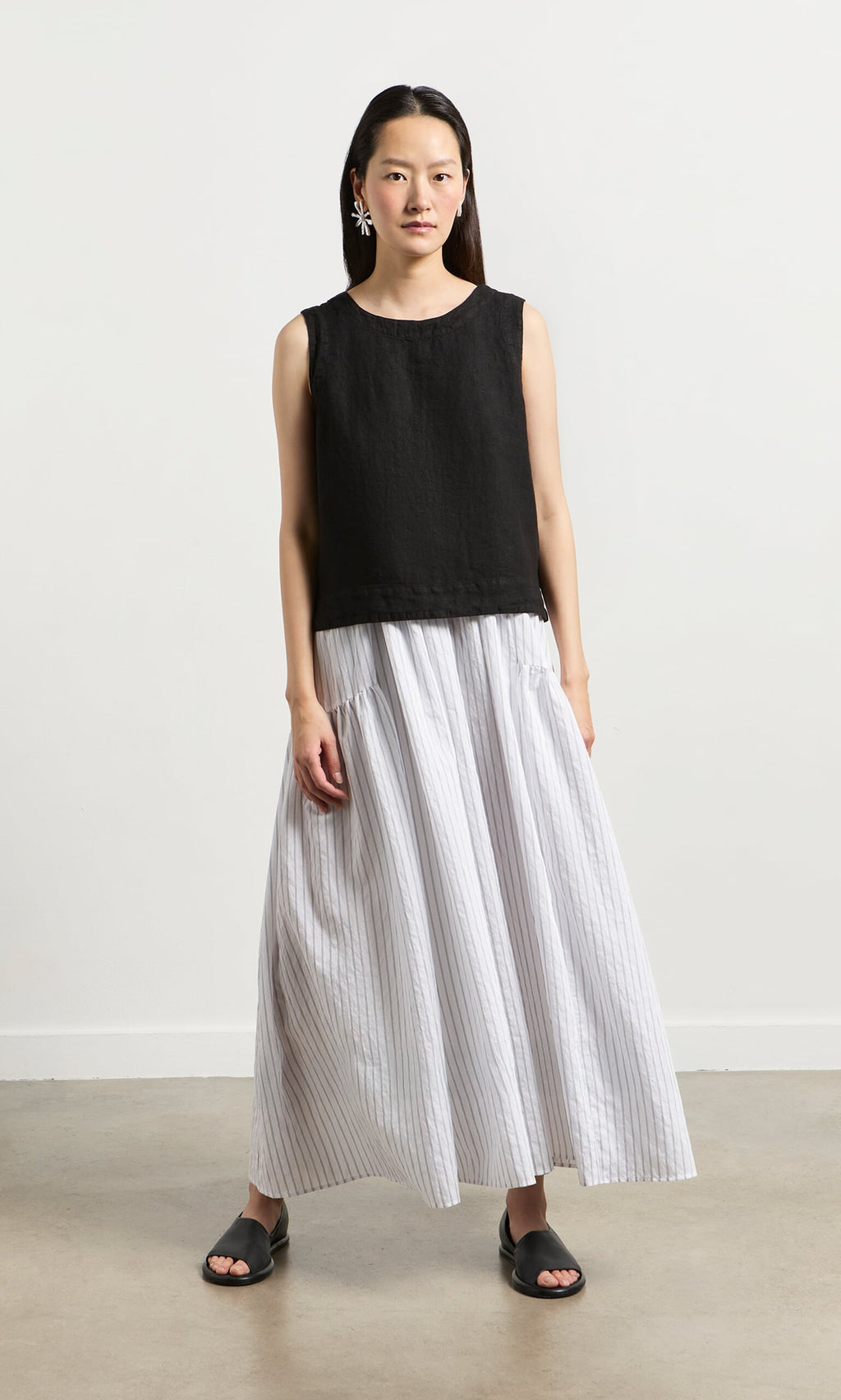 Plümo- A woman in the Linda Top - Solid Black, a white striped skirt, and pink earrings stands on a plain floor in San Francisco.