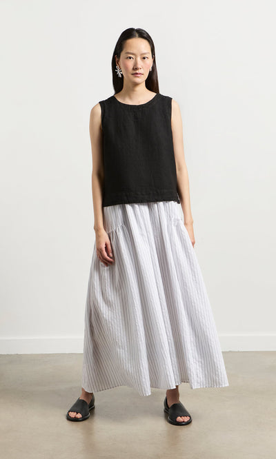 Plümo- A woman in the Linda Top - Solid Black, a white striped skirt, and pink earrings stands on a plain floor in San Francisco.