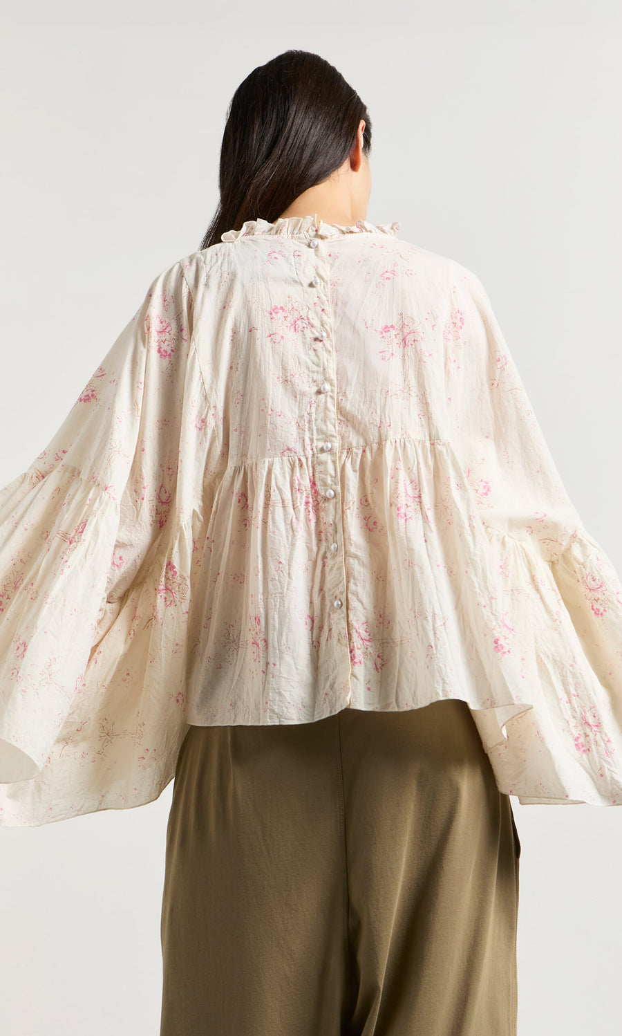 Plümo- A woman facing away in a Litchen Blouse with vintage floral print, ruffles, back buttons, and olive green pants.