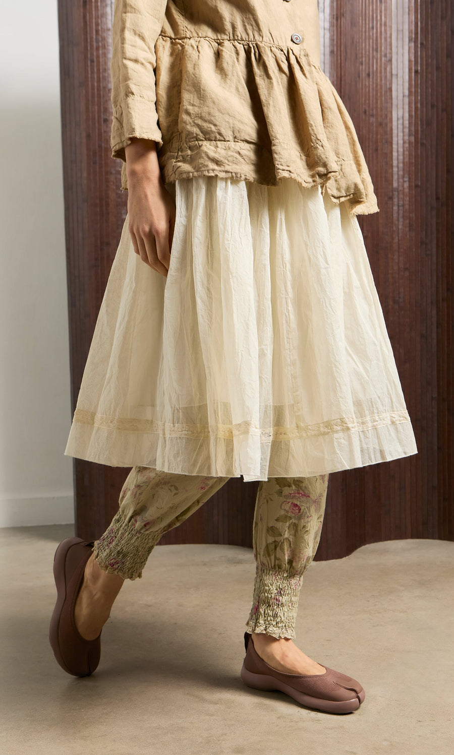 Plümo- Person wearing a beige tunic, Lou Organza Skirt, floral leggings, and brown shoes on a concrete floor.