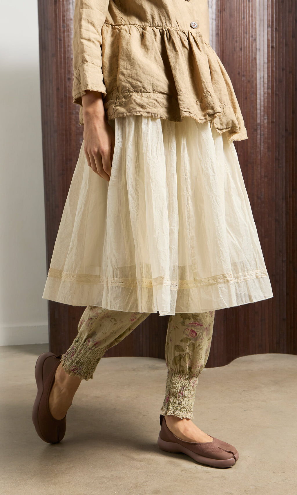 Plümo- Woman stands indoors on a concrete floor wearing Choco Tabi Ballerinas with a beige tunic, white skirt, and floral leggings.
