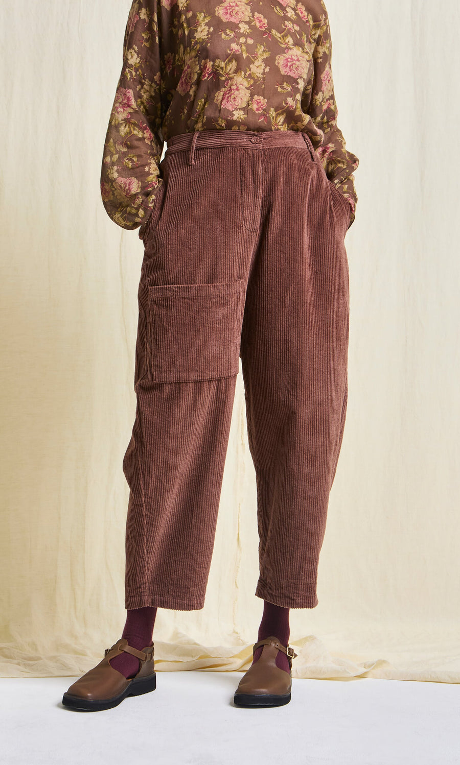 Plümo- Person in high waist Lumber Pants, a floral sheer blouse, maroon socks, and brown shoes stands against a light background.