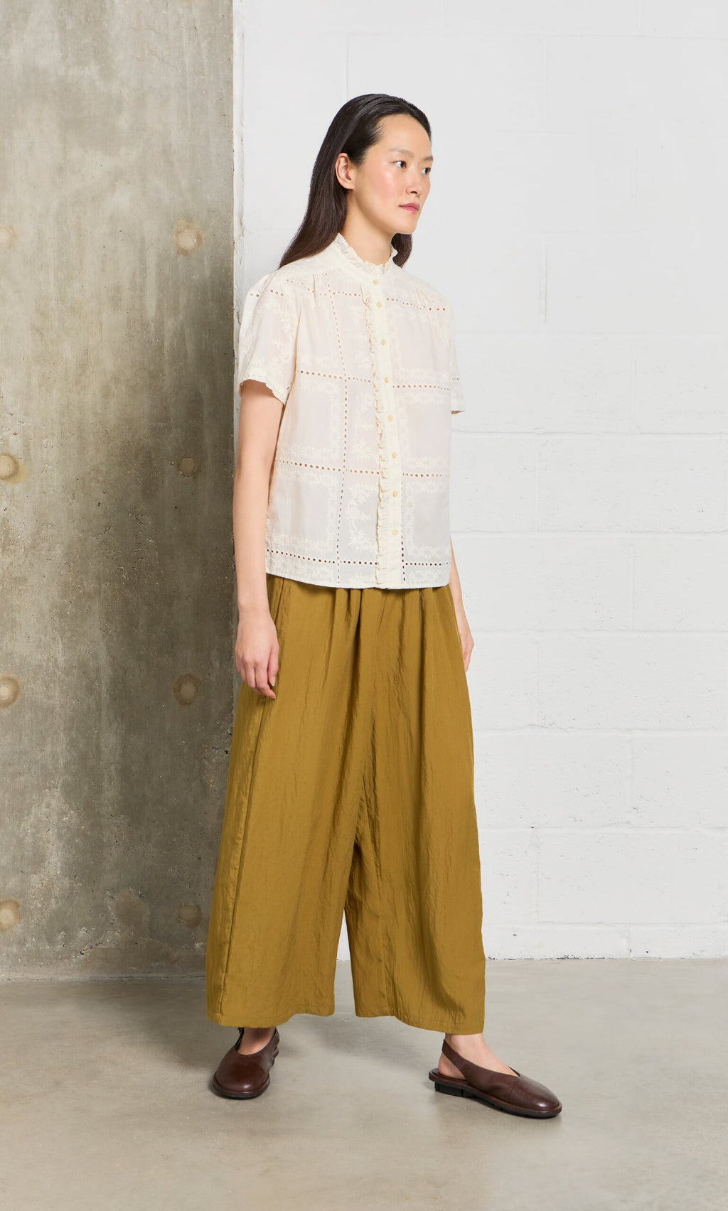 Plümo- Woman wearing the Gill Blouse and wide mustard pants stands against a concrete and white wall.